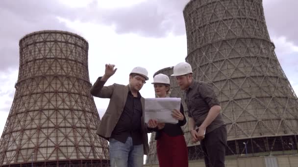 Grupo de arquitetos discutindo um plano de construção no contexto da torre de resfriamento da usina nuclear. Usina a carvão. designers em capacetes de construção . — Vídeo de Stock