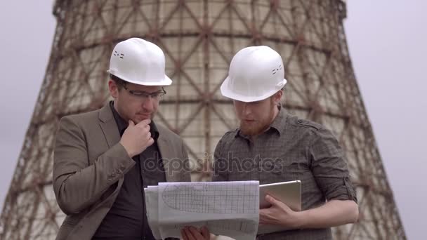 Zwei Geschäftsleute mit Bauhelmen auf dem Hintergrund des Atomkraftwerks. Architekten im Gespräch mit einem Bauplan — Stockvideo