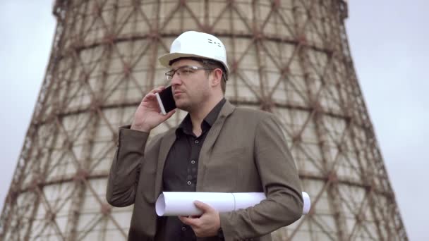発電所の背景に若い建築家。魅力的な男性のデザイナーの図面を押しながら携帯電話で話しています。原子力発電所の塔を冷却します。石炭燃焼発電所 — ストック動画