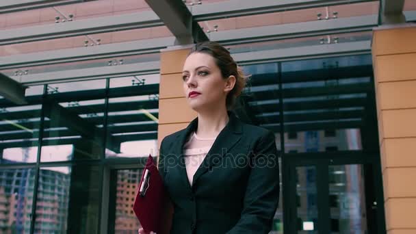 Gyönyörű magabiztos fiatal üzleti nő a háttérben az a modern üzletközpontot is kínál. Lány üzleti ruhákat, egy piros mappát, kezében a háttérben egy irodaház. Lassú mozgás — Stock videók
