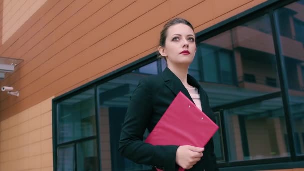 Die schöne selbstbewusste junge Geschäftsfrau hat es eilig zu verhandeln. Mädchen in Businesskleidung mit einem roten Ordner in der Hand vor dem Hintergrund eines Bürogebäudes. Zeitlupe — Stockvideo