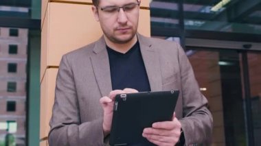 Bir tablet bilgisayarda çalışan emin iş adamı. Adam bir ceket ve modern iş binanın arka planda gözlük. Elinde akıllı izlemek ve tablet bilgisayar.