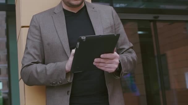 Уверенный бизнесмен работает за планшетным компьютером. Человек в пиджаке и очках на фоне современного бизнес-здания. Умные часы и планшетный компьютер в руках . — стоковое видео