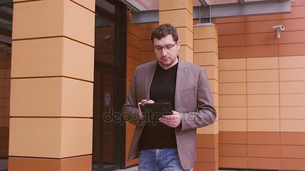 Hombre de negocios moderno caminando cerca del centro de negocios y trabajando en la tableta. El hombre mira un reloj inteligente. Hombre con chaqueta y gafas en el fondo de un moderno edificio de negocios . — Vídeos de Stock