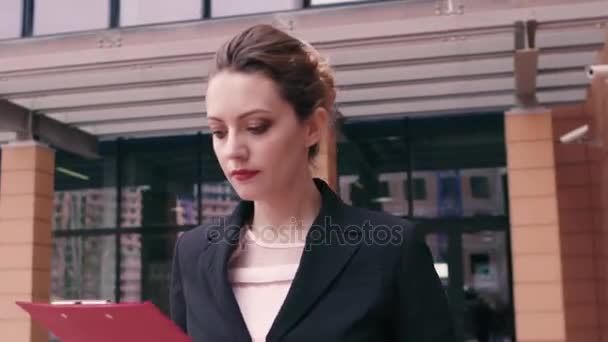 Bella fiduciosa giovane donna d'affari ha fretta di negoziare e guarda l'orologio. Ragazza in abiti da lavoro con una cartella rossa in mano sullo sfondo di un edificio per uffici — Video Stock