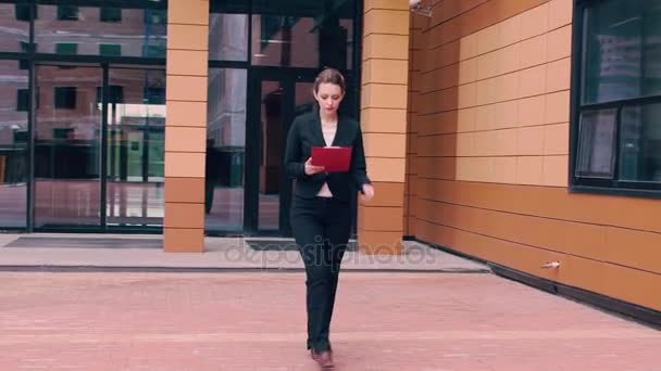 Beautiful confident young business woman is in a hurry to negotiate and looks at the clock. Girl in business clothes with a red folder in hands on the background of an office building — Stock Video
