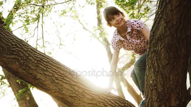 Счастливый ребенок забрался на деревья. Девушка сидит на ветке дерева и улыбается. Малыш играет в парке. замедленное движение — стоковое видео