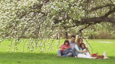 Baba, anne ve çocuk bir çayır çiçek açması bir elma ağacının altında oturuyor. Mutlu aile doğada dinleniyor. İki küçük kız onların anne kucağına oturuyor