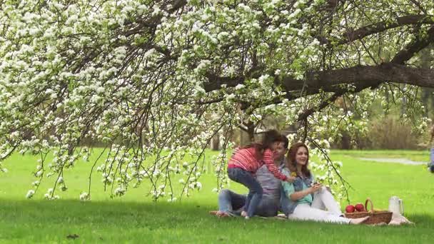 Szczęśliwa rodzina spędza czas na piknik w pobliżu kwitnących jabłoni. Tata, Mama i dwie siostry spędzają czas odkryty. Dzieci dają rodzicom kwiaty na łące — Wideo stockowe