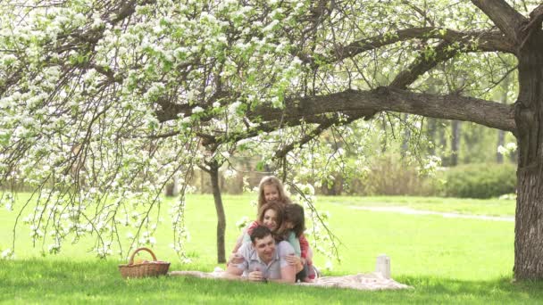 Fröhliches Porträt einer jungen Familie. Eltern liegen mit ihren Kindern unter einem blühenden Apfelbaum. Familie hat Spaß beim Picknick — Stockvideo