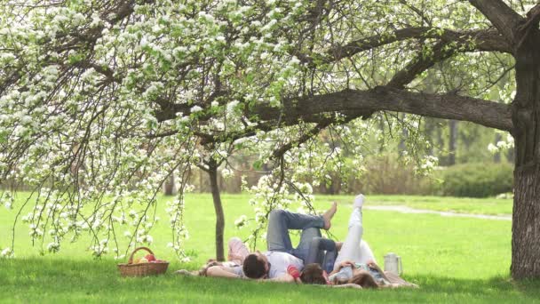 Ritratto allegro di una giovane famiglia. I genitori con i loro figli giacciono sotto un melo in fiore. Famiglia divertendosi al picnic — Video Stock