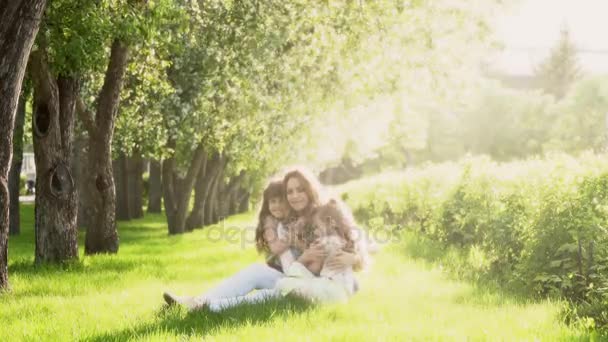 Mamá y dos hijas sentadas en la hierba cerca de los manzanos. niños y madre en el Parque al atardecer. la relación de licitación entre madre e hija . — Vídeo de stock