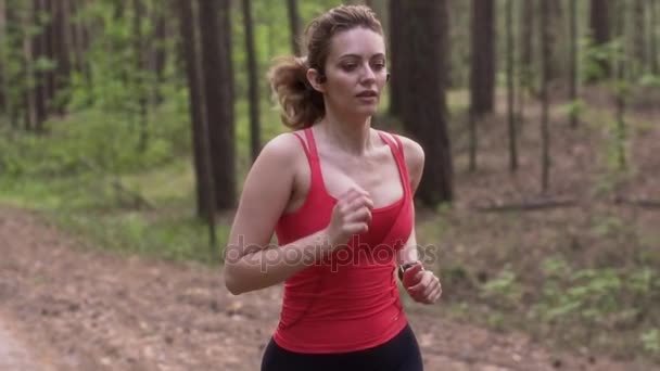 Joven atractiva mujer corriendo en un bosque de verano. chica con auriculares inalámbricos y un reloj con monitor de frecuencia cardíaca deportes. Entrenamiento de protectores. cámara lenta — Vídeos de Stock