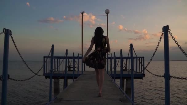 Silhouet van een mooi meisje in een lichte jurk bij zonsondergang. Een meisje loopt een pier in de zee — Stockvideo