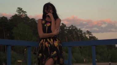 Gün batımı kameraya poz ve gülümseyen güzel genç kız. Gün batımında ışık elbiseli güzel bir kız silüeti. Bir kız bir iskele deniz üzerinde yürüyüş