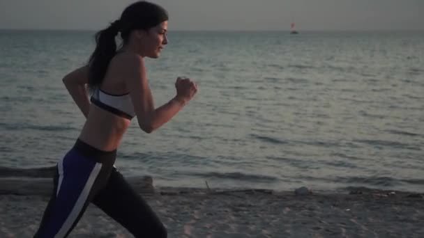 Retrato de una deportista corriendo por la playa al atardecer. Chica de 20 años corriendo en la arena en la playa al atardecer. Correr al aire libre. cámara lenta — Vídeos de Stock