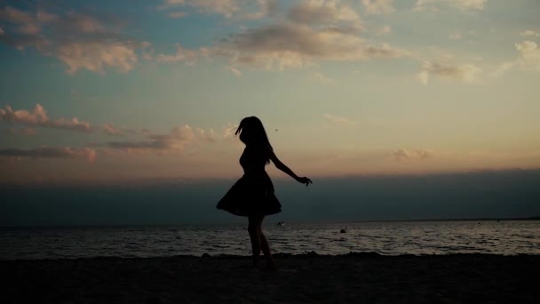Szép, boldog fiatal lány örvény a parton és boldogan integet a karját. Sziluett naplementekor. Fiatal nő napnyugtakor a tengerparton. lassú mozgás — Stock videók