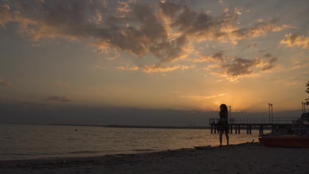 Σιλουέτα ενός κοριτσιού σε ένα ελαφρύ φόρεμα στην παραλία το ηλιοβασίλεμα. Όμορφη νεαρή κοπέλα περπάτημα στην άμμο ξυπόλυτη — Αρχείο Βίντεο