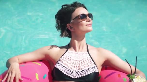 Jeune femme en lunettes de soleil buvant un cocktail et bronzant couché sur une martrase gonflable dans la piscine — Video