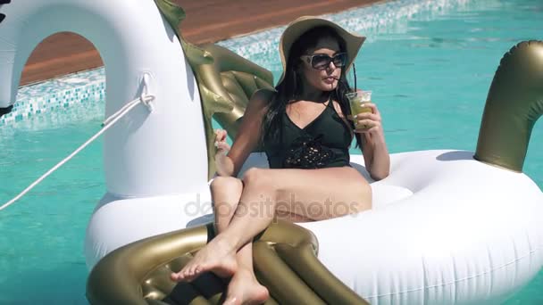 Hot girl sexy in bikini beve cocktail sdraiato su materasso gonfiabile pegasus in piscina all'aperto. Giovane donna in occhiali da sole e un cappello di paglia prendere il sole sulla spiaggia — Video Stock