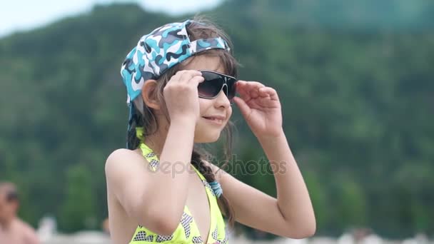 Niña en gafas de sol y traje de baño sonriendo y posando para la cámara. niño de vacaciones junto al agua. Movimiento lento — Vídeo de stock