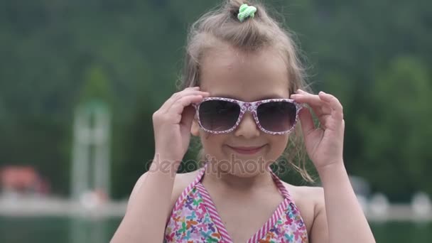 Маленькая красивая девочка в купальнике и солнцезащитных очках улыбается и позирует перед камерой. Медленное движение — стоковое видео