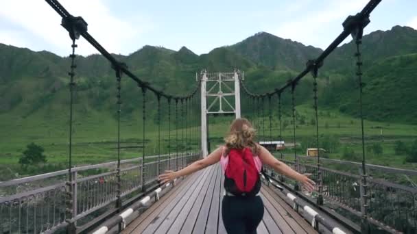 Девушка-путешественница с рюкзаком бежит вдоль моста. туристка на фоне гор. замедленное движение — стоковое видео