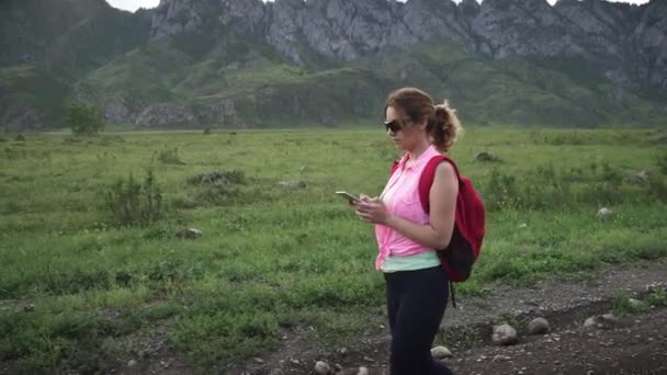 Toeristische meisje loopt langs een onverharde weg tegen de achtergrond van de bergen. Vrouw reiziger met een rugzak. toerist met een mobiele telefoon in zijn handen signaal een — Stockvideo