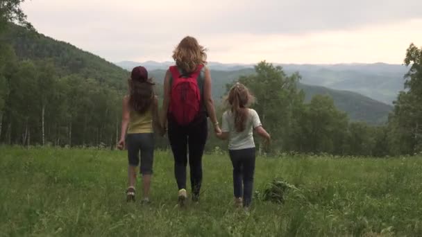 क्रीडा कुटुंब पर्वतावरुन प्रवास करते. जंगलाच्या ग्लेडद्वारे आपल्या मुलांना अग्रगण्य बॅकपॅकसह पर्यटकाची एक तरुण आई — स्टॉक व्हिडिओ