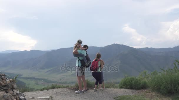 Сім'я туристів. Батьки тримають своїх дітей на руках і показують їм прекрасний вид на гори. Мандрівники з маленькими дітьми в горах — стокове відео