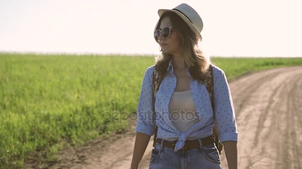 Lány utas visel, napszemüveget és séta lefelé az úton, a mező kalapot. fiatal nő túrázó a hátizsák mentem kemping. lassú mozgás — Stock videók