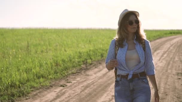 Fille hipster dans un chapeau et des lunettes de soleil marche sur la route d'une prairie — Video