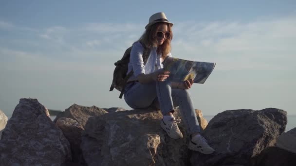 Lány turisztikai holding megjelenítése a kezében, és álmodik az új utazik. fiatal nő ül a sziklákon nyugszik — Stock videók