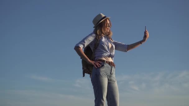 Kız Gezgin selfie cep telefonu kamera yapar. Genç kadın bir sırt çantası ile bir turtsey var. Kız güneş gözlüğü ve şapka gökyüzü ve bulutlar karşı — Stok video