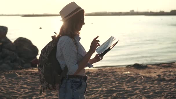 Mädchen ist eine Touristin wählt ein Reiseziel auf der Landkarte. junge Hipsterin mit Sonnenbrille und Hut. Mädchen bei Sonnenuntergang am Strand — Stockvideo