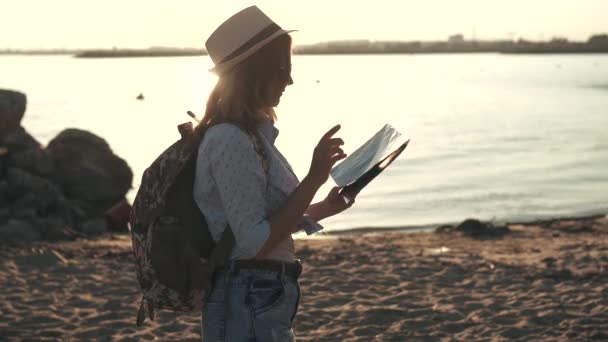 Girl es un turista elige un destino turístico en el mapa. mujer joven hipster con gafas de sol y un sombrero. chica al atardecer en la playa — Vídeos de Stock