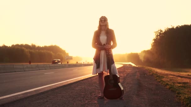 Mädchen-Hipster mit Gitarre trampen bei Sonnenuntergang auf der Straße. Porträt eines jungen schönen Mädchens mit Sonnenbrille. Mädchen-Hipster im Morgengrauen — Stockvideo