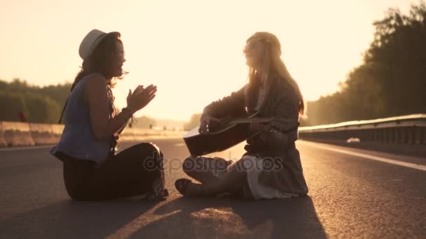 As raparigas hippies cantam canções e tu tocas guitarra. dois hipsters sentados na estrada ao pôr-do-sol — Vídeo de Stock