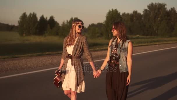İki hippi kız el ele tutuşup yolda yürüyor. Elinde gitar olan genç bir kadın ve gün batımında şapka ve güneş gözlüklü arkadaşı. — Stok video