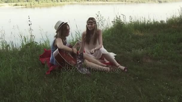 Girls in hippie clothes play guitar and sing while sitting on the cliff near the river — Stock Video