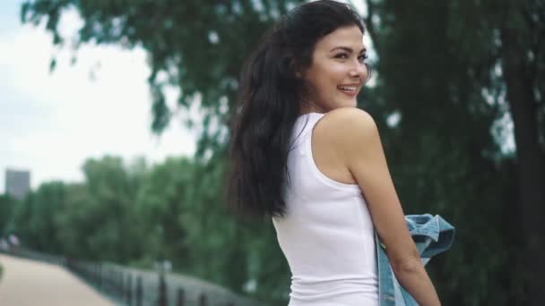 Красивая молодая девушка улыбается и позирует на камеру. Портрет привлекательной девушки в белом топе танка на фоне зеленых деревьев. замедленное движение — стоковое видео