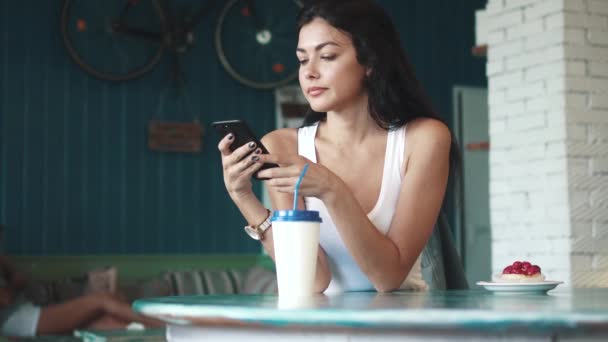 Sexy chica disfruta de un teléfono inteligente y bebe café sentado en un acogedor café — Vídeos de Stock