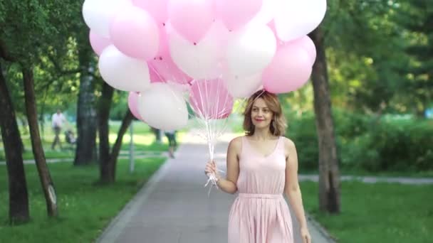 Hermosa joven felizmente va en un parque de verano sosteniendo globos . — Vídeo de stock