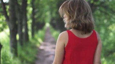 Kırmızı elbiseli bir Linden sokak yürüyüş bir genç kadın. Kız uzakta kameradan yürür ve şakacı etrafına bakıyor. ağır çekim