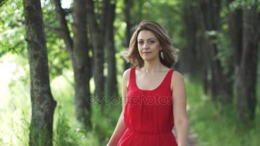 Kırmızı elbiseli güzel bir genç kadının duygusal portre. Kız yeşil sokak yürüyüş ve gülümseyerek. ağır çekim