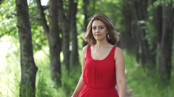 Sensuele portret van een mooie jonge vrouw in een rode jurk. Het meisje loopt langs de groene steeg en glimlachen. Slow motion — Stockvideo