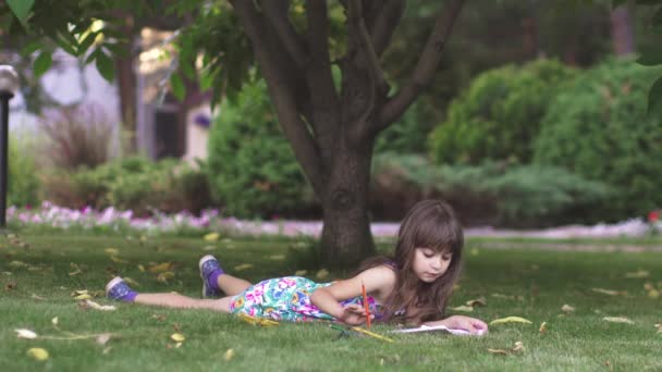 Niño pintar acostado en la hierba debajo de un árbol. niña dibujar con lápices de colores en el patio trasero — Vídeos de Stock