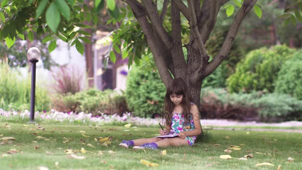 키즈 페인트 나무 아래 잔디에 앉아. 어린 소녀는 뒤뜰에 크레용으로 그려 — 비디오