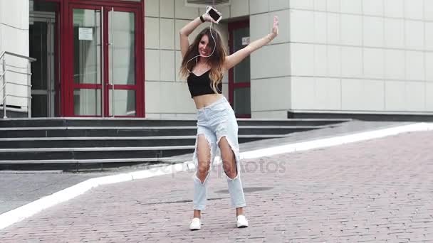 Chica joven es divertido bailar en la calle y escuchar música en los auriculares. Hermosa chica en pantalones vaqueros andrajosos y un chaleco se divierte. cámara lenta — Vídeo de stock