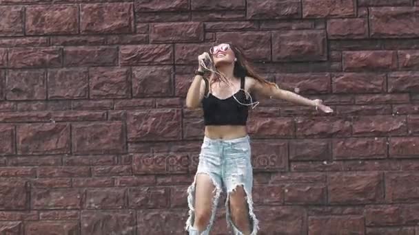 En våt tjej sjunger låtar i en mobiltelefon istället för en mikrofon och danser som är galen under spray vatten på gatan. slowmotion — Stockvideo